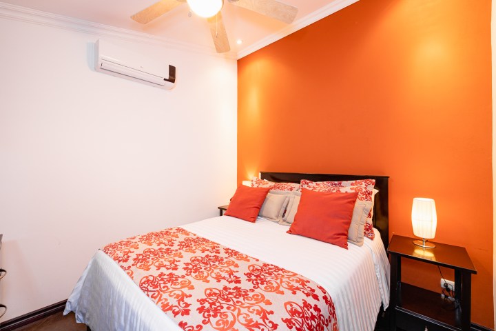 Bedroom with orange accent wall, queen bed, red pillows, bedside lamps, ceiling fan, and air conditioning.