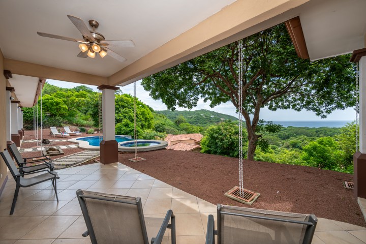 Covered patio with chairs, ceiling fan, overlooking a pool and lush green landscape with distant ocean view.