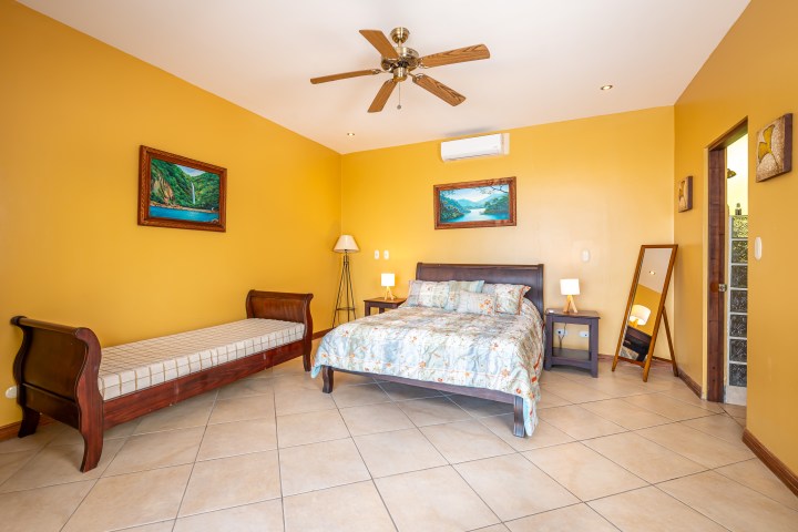 Spacious bedroom with yellow walls, bed, bench, paintings, mirror, and ceiling fan.
