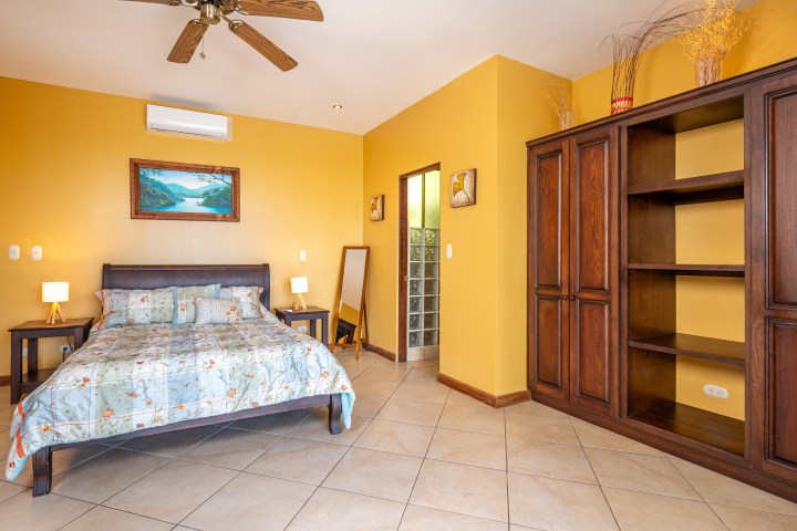 Bedroom with yellow walls, bed with floral bedding, wooden shelves, and a ceiling fan.