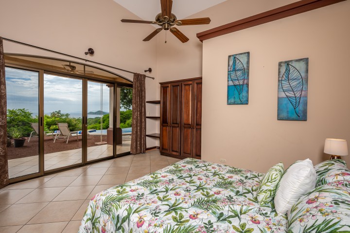 Bedroom with floral bedspread, ceiling fan, art on walls, and glass doors opening to a patio with chairs and greenery.
