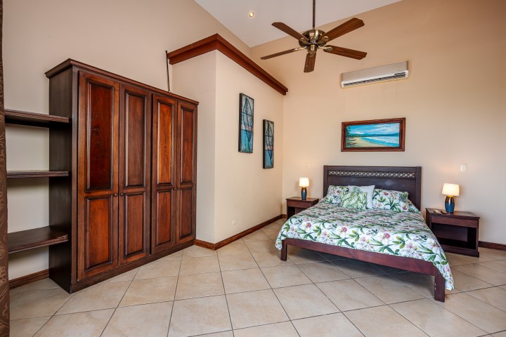 Bedroom with a wooden wardrobe, floral bedspread, ceiling fan, and two paintings on cream walls.