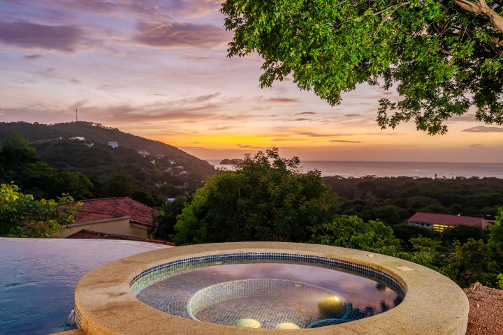 Round hot tub overlooking sunset view of hills and ocean with trees.