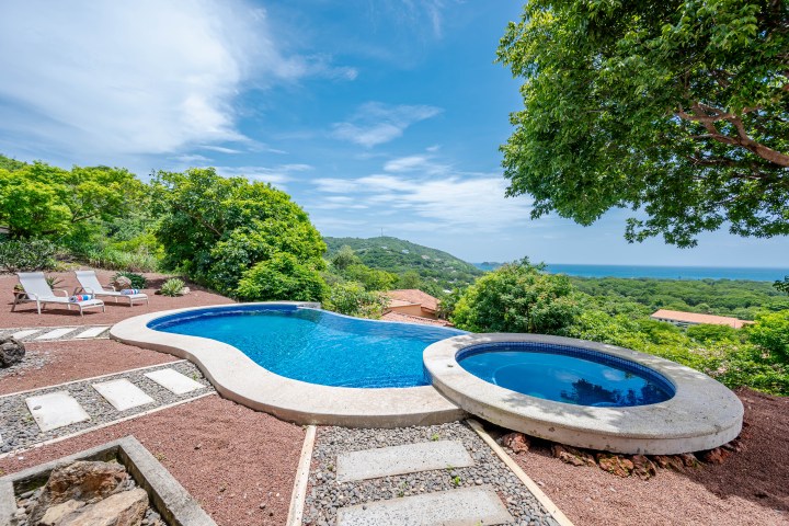 Curved pool and jacuzzi with ocean view, surrounded by greenery and lounge chairs.