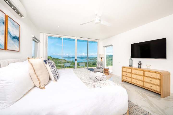 Bright bedroom with ocean view, white bedding, and wooden dresser, featuring a large window and ceiling fan.