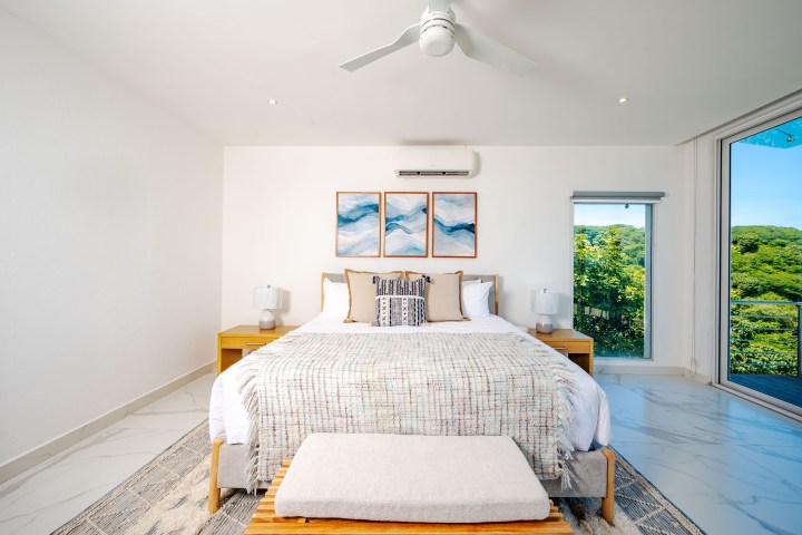 Modern bedroom with white bed, artwork above, lamps, and large window with scenic view.