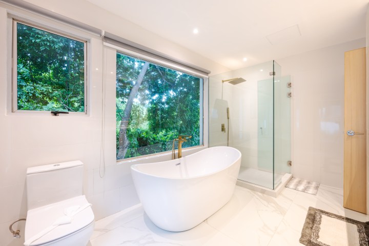 Modern bathroom with freestanding tub, glass shower, large window with green view, and white fixtures.