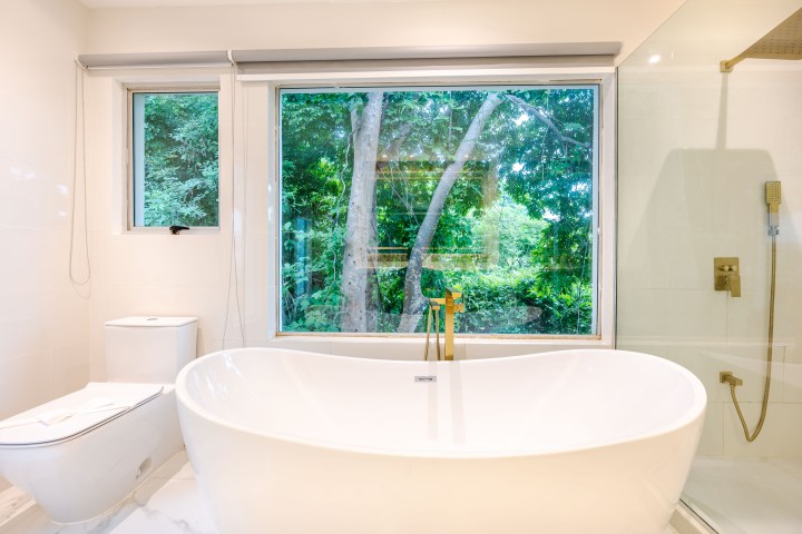 Modern bathroom with freestanding tub, glass shower, and large window overlooking greenery.