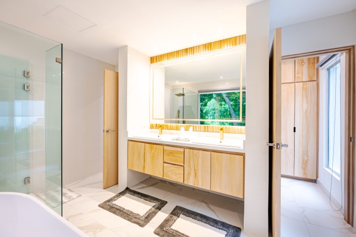 Modern bathroom with wooden vanity, large mirror, glass shower, and open doorway to a bright room.