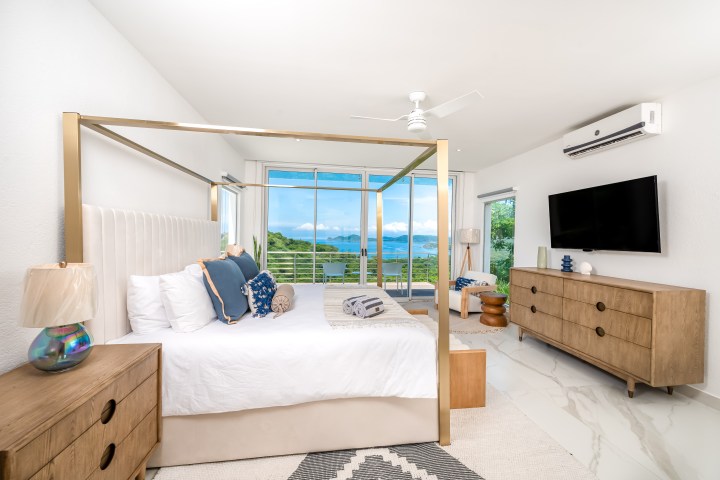 Modern bedroom with canopy bed, dresser, TV, and large window view of greenery and water.