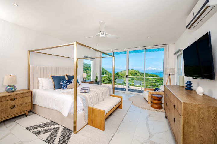 Modern bedroom with a canopy bed, wooden furniture, and a balcony view of greenery and ocean.