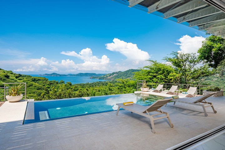 Infinity pool with loungers overlooking ocean and hills under a clear blue sky.