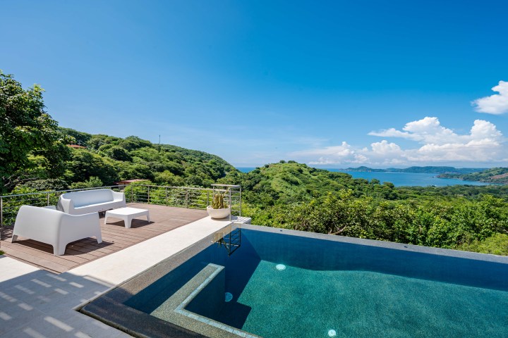 Terrace with seating, pool, and lush landscape overlooking distant blue ocean and sky.