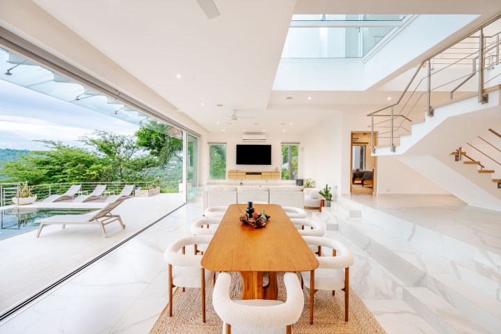 Modern open-plan living room with dining table, white chairs, and sliding doors to a patio with lounge chairs.