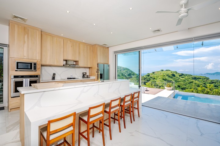 Modern kitchen with wood cabinets, island seating, and view of pool and lush landscape through large windows.