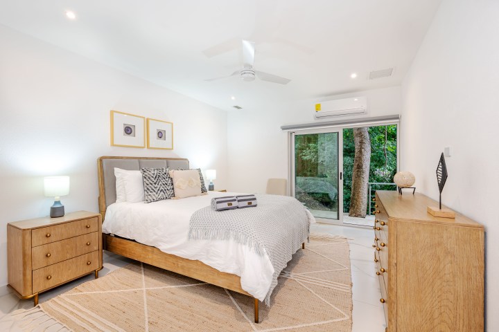 Modern bedroom with a large bed, wooden furniture, and a sliding glass door overlooking trees.