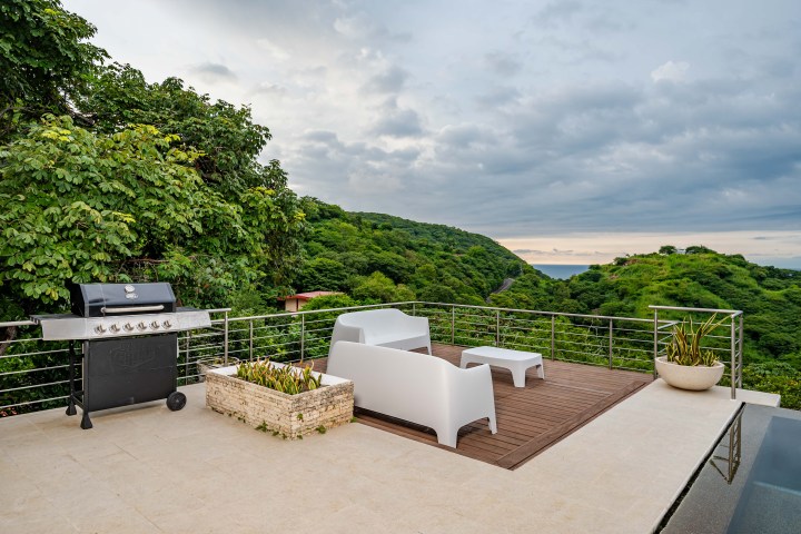 Rooftop patio with white seating, grill, lush green hills, and ocean view at sunset.