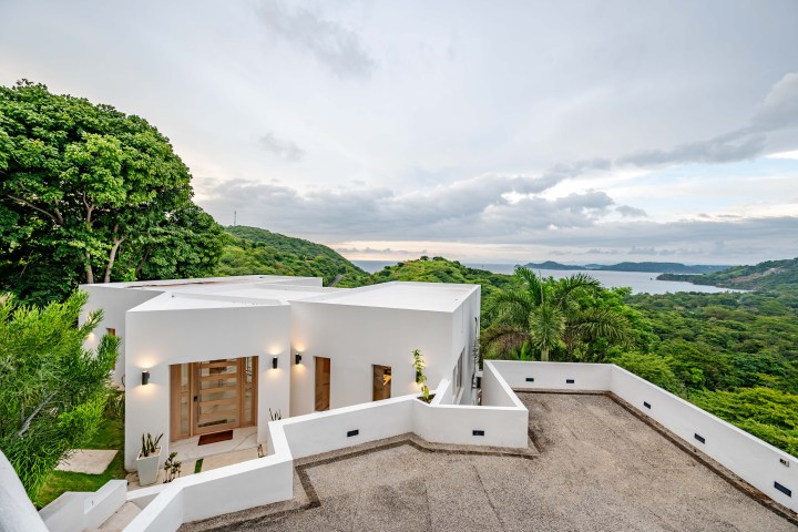 Modern white house with a scenic ocean view, surrounded by lush greenery.