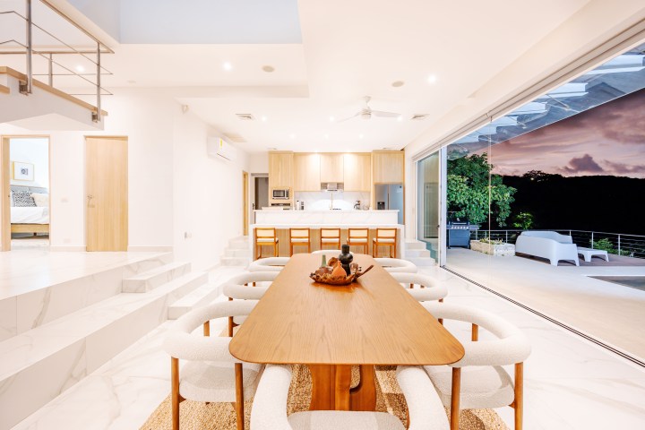 Modern, open-plan kitchen and dining area with large glass doors opening to a patio and a view of trees at sunset.