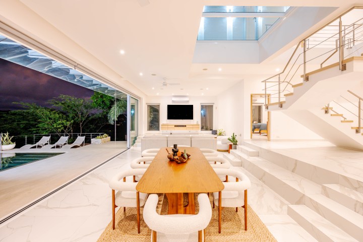 Modern open living space, dining area with wooden table, stairs, poolside view, and neutral tones.