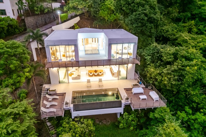 Modern white house with pool surrounded by lush green trees, photographed from above.