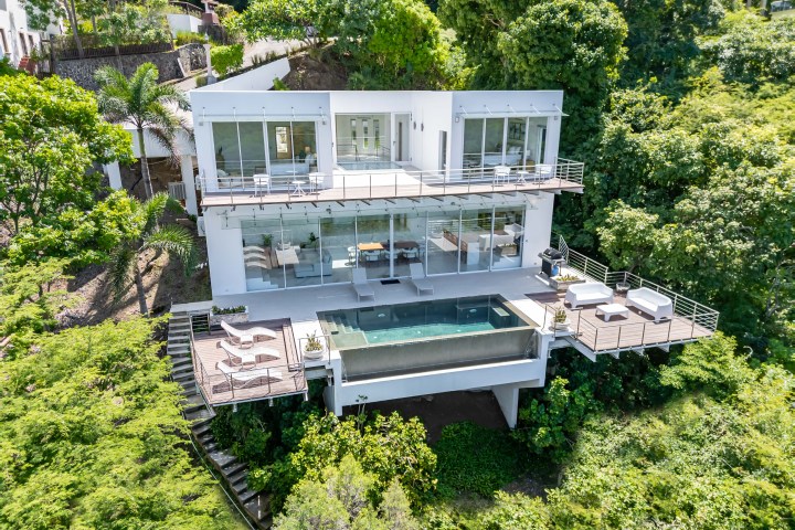Modern hillside house with glass facade, terraces, and pool surrounded by lush greenery.