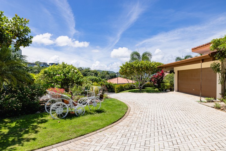 House with a brick driveway, landscaped garden, and a decorative white cart under a blue sky.