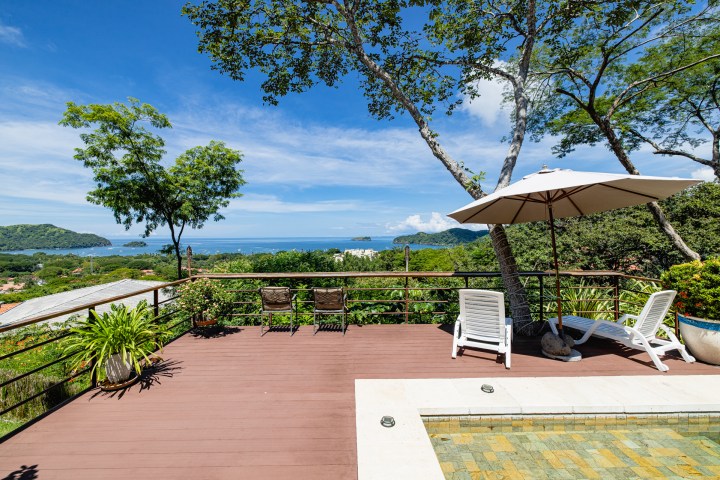 Deck with lounge chairs and umbrella overlooking a scenic view of the ocean and green hills.