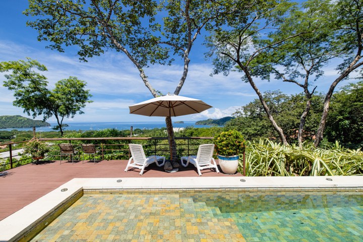 Terrace with pool, loungers, umbrella; overlooking trees and ocean under a blue sky.