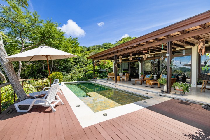 Modern patio with pool, sun loungers, umbrella, and greenery; glass doors lead to deck with seating areas.