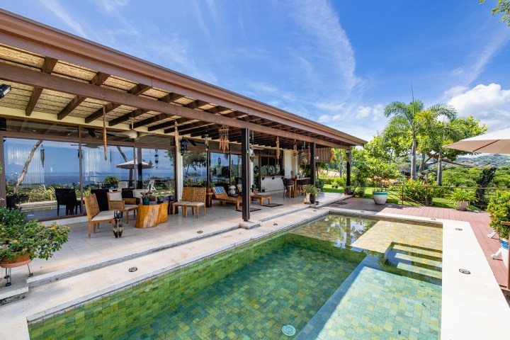 Open patio with seating area overlooking a pool and garden under a clear sky.