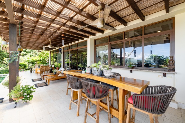 Covered patio with wooden furniture, plants, and ceiling fans overlooking a green garden.