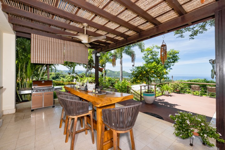 Outdoor patio with wooden table, chairs, ceiling fan, and ocean view in the background.