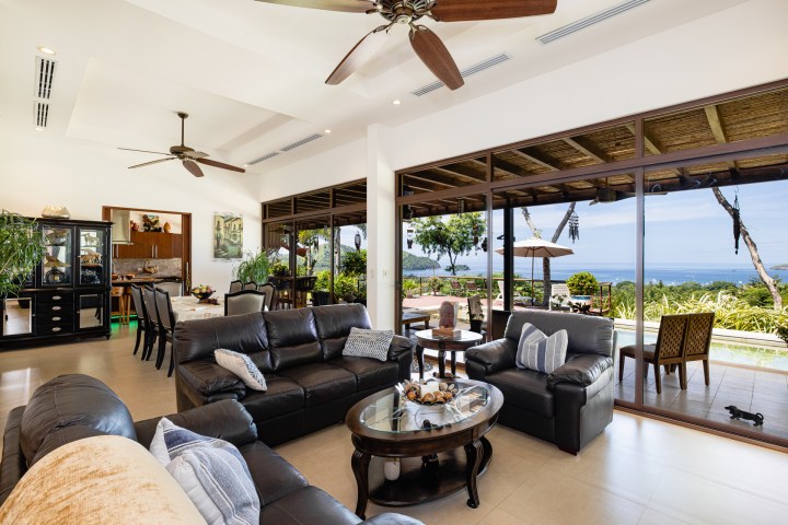 Luxurious living room with leather sofas, ceiling fans, sea view through large windows, and outdoor patio.