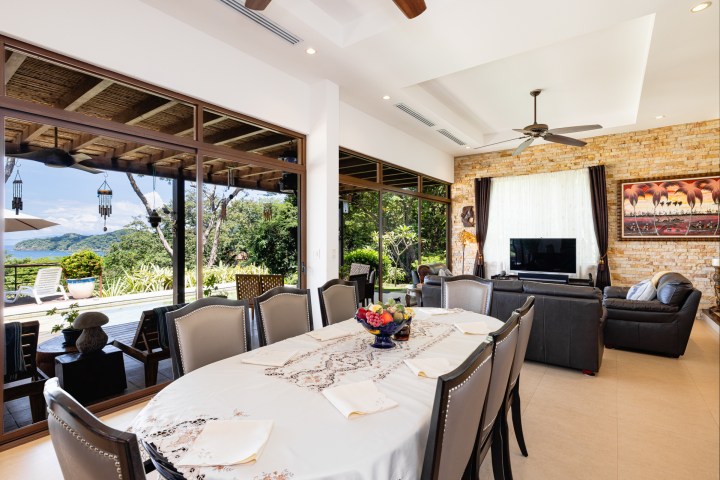 Modern living room with dining table, couches, TV, and large windows overlooking a garden and hills.