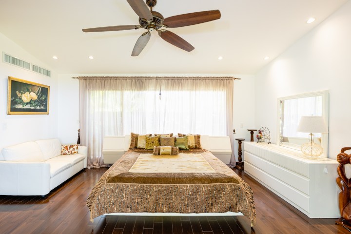 Bright bedroom with a large bed, ceiling fan, white sofa, dresser, and large window.