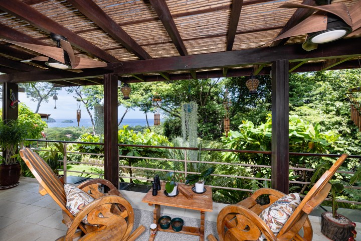Outdoor patio with wooden chairs, table, and lush green garden overlooking the ocean.