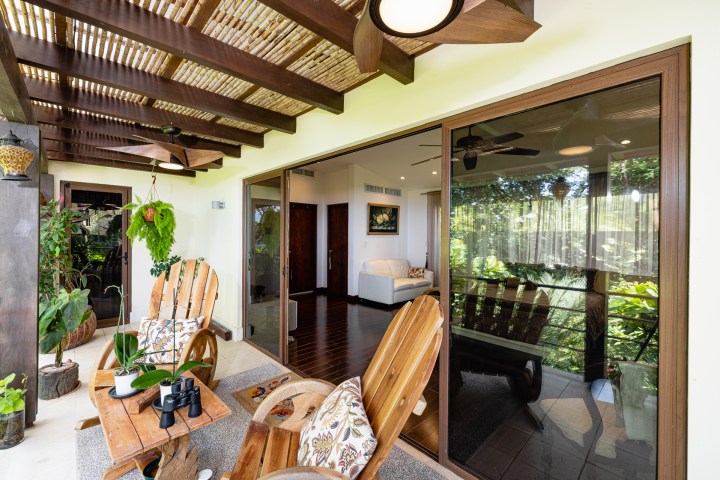 Cozy patio with wooden chairs, plants, and a view into a living room through glass doors.