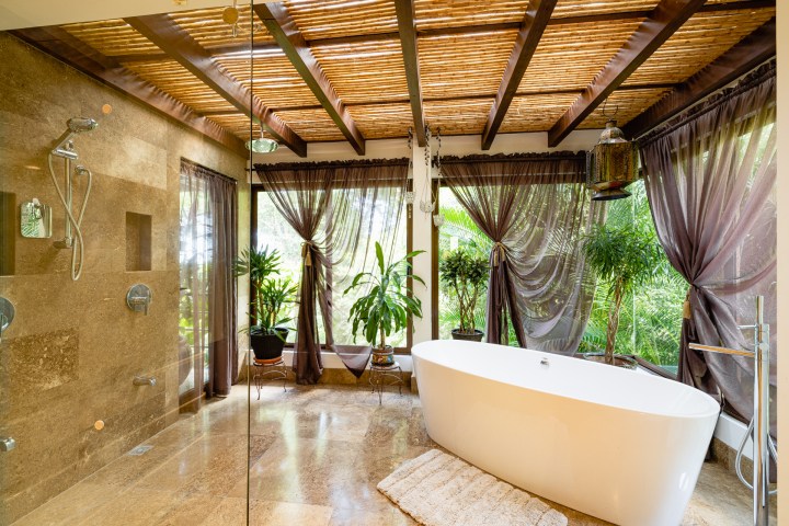 Luxurious bathroom with freestanding tub, glass shower, and plants under a bamboo ceiling with sheer curtains.