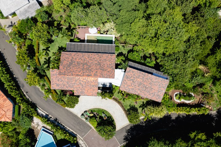 Aerial view of a house with a red-tiled roof, surrounded by greenery and a circular driveway.