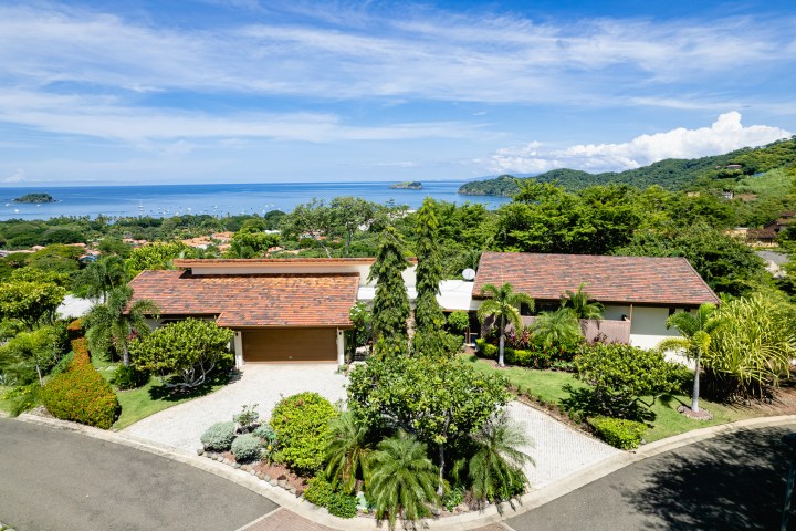 House with a red roof and lush garden overlooking a bay with boats and green hills.