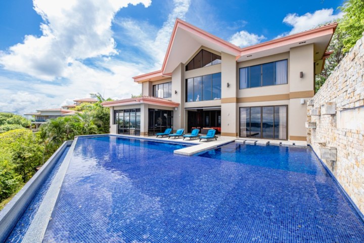 Modern villa with large infinity pool, blue loungers, and palm trees under a cloudy sky.