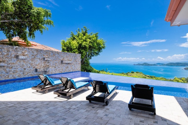 Infinity pool with lounge chairs overlooking ocean and islands under a clear blue sky.