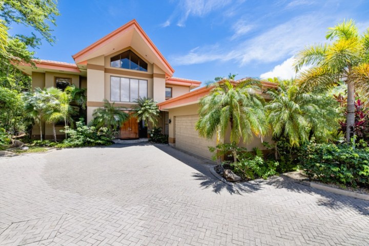 Large house with orange roof, palm trees, and paved driveway under a blue sky.