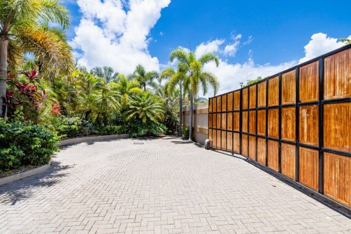 Paved driveway surrounded by palm trees, wooden fence, and clear blue sky.