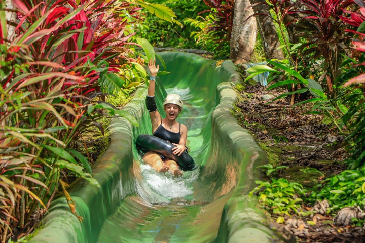 Person in helmet tubing down a green water slide amidst lush tropical foliage.