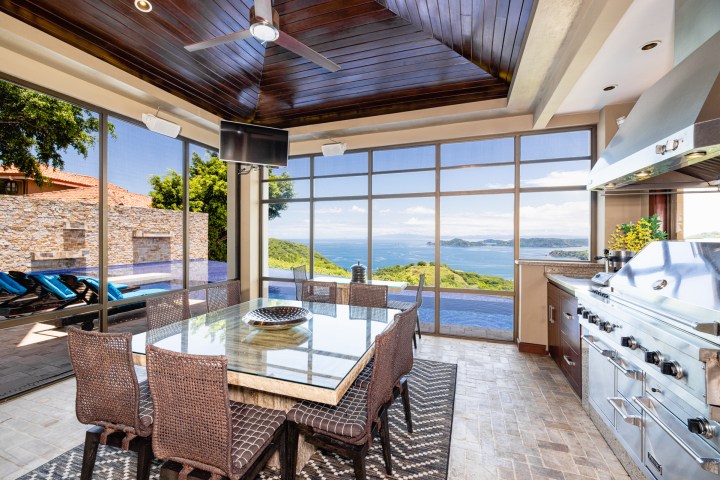 Luxury outdoor kitchen with dining table, barbecue grill, and ocean view through large windows.