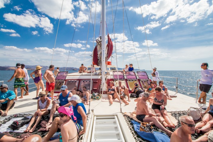 People relaxing on a sailboat under a bright blue sky with clouds and distant land visible.