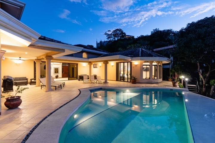 Elegant villa with illuminated pool and patio furniture at dusk, surrounded by trees.