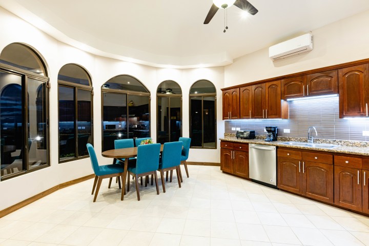 Spacious kitchen with curved windows, wooden cabinets, and a round table with blue chairs.
